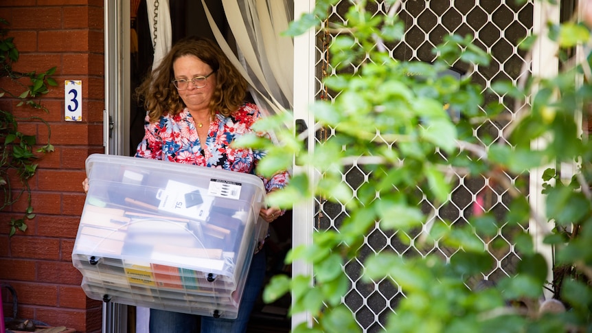 A woman wearing a bright flowery blouse carries a box of things outside her home. 