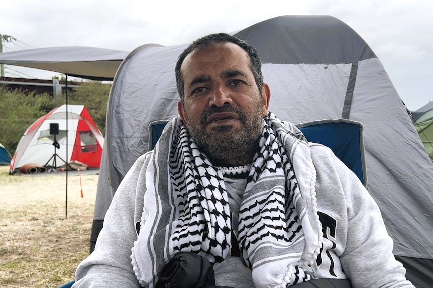 A man sits in a chair, a palestinian scarf over his shoulders. Tens in view in the background.