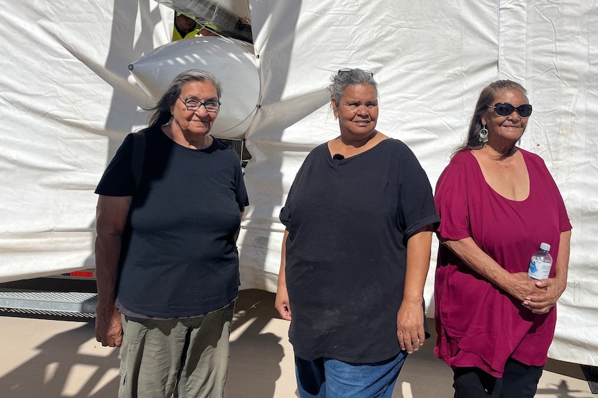 three Indigenous women stand in front of a white plastic shelter with rocket nose poking out