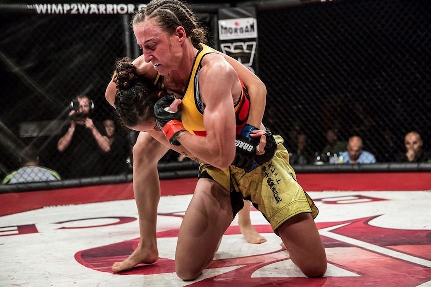 A woman kneeling with another women in a headlock inside a mixed martial arts cage