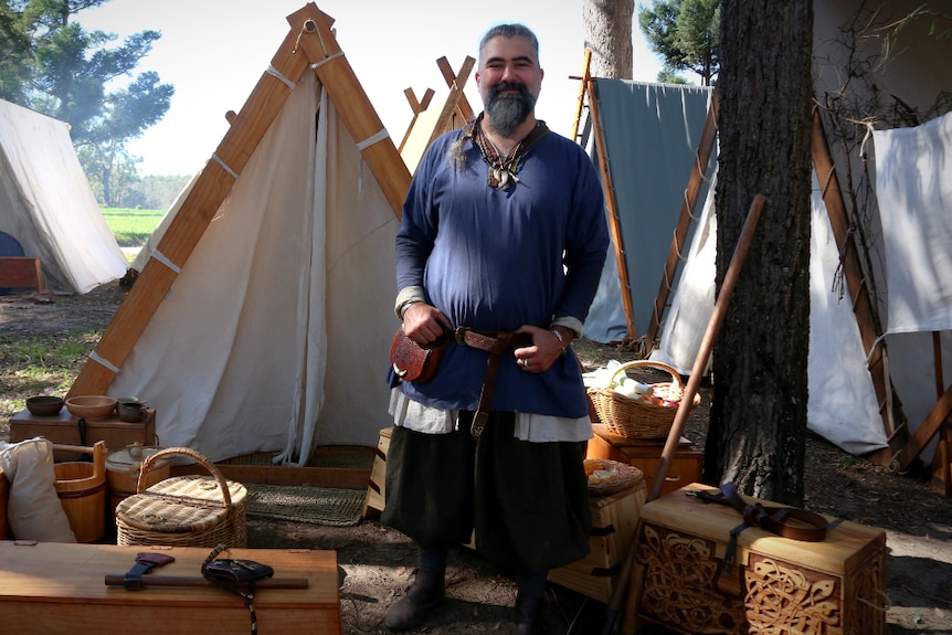 A man dressed in Viking clothes stands in front of an A-frame tent.