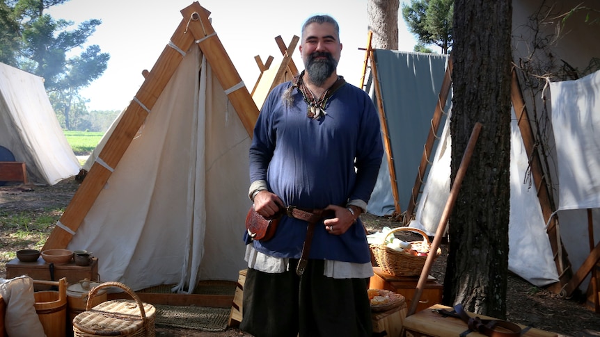 A man dressed in Viking clothes stands in front of an A-frame tent.
