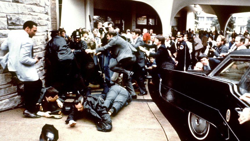 Secret service agents cover presidential staff as a chaotic crowd forms at the scene of the attempted assassination.