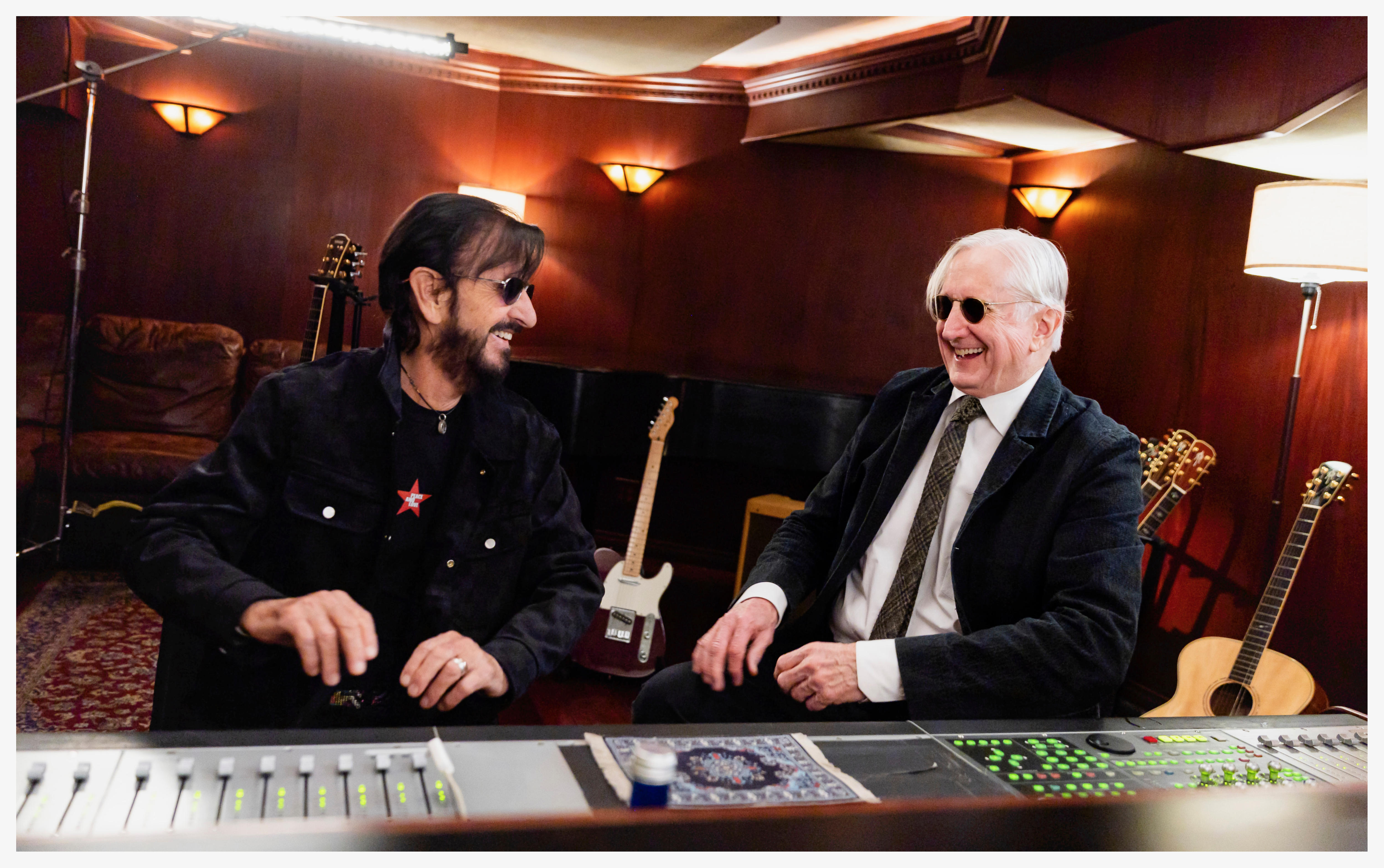 Ringo Starr y T Bone Burnett se sientan frente a una mesa de mezclas en un estudio de grabación con paneles de madera. Ambos ríen.