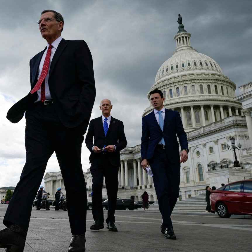 A group of men walk along a street in front of the US Capitol on a stormy day.
