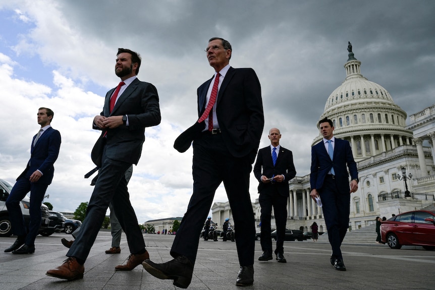 A group of men walk along a street in front of the US Capitol on a stormy day.