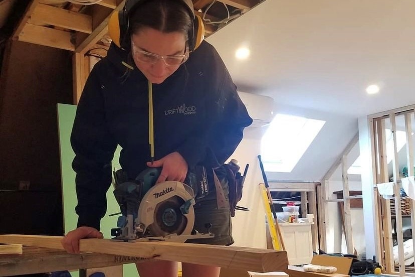 A woman in work wear and ear protection using a power tool.