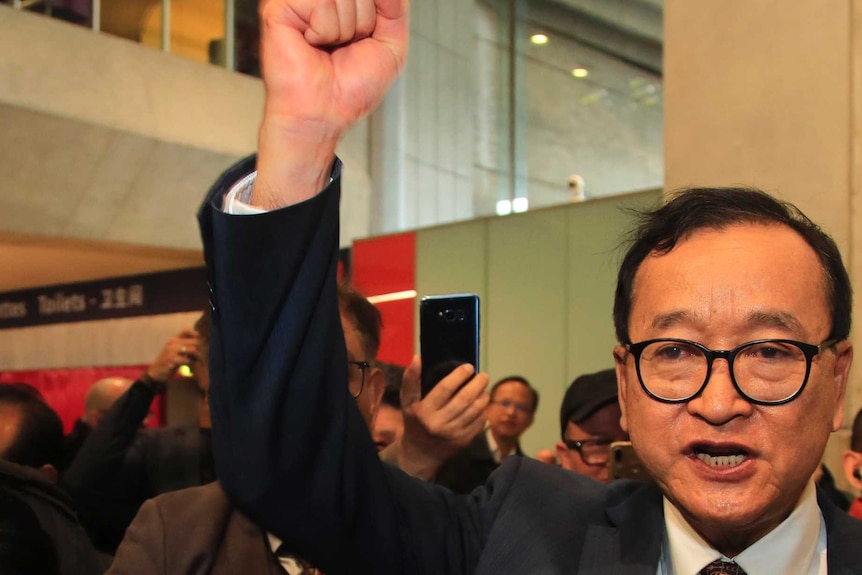 Man wearing round glasses, suit and tie, and surrounded by men and women supporters, raises fist in the air and shouts slogans