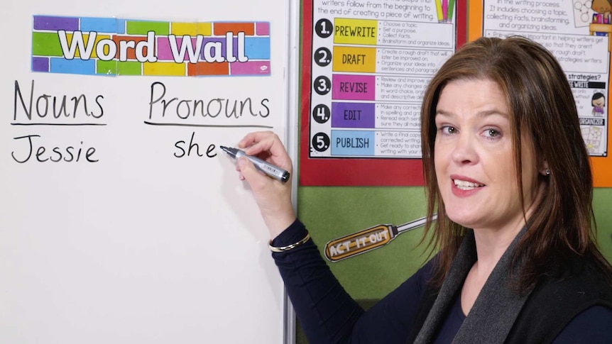 Female teacher writes on whiteboard, contains words "nouns", "pronouns"