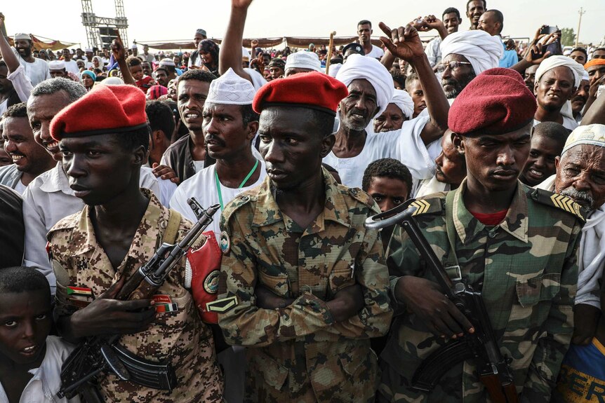Three men in military camouflage and red berets stand holding weapons in front of a crowd of men wearing white