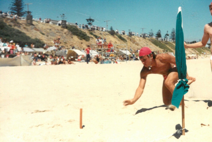 A boy in a lifesaving hat leaps across the sand towards a small post