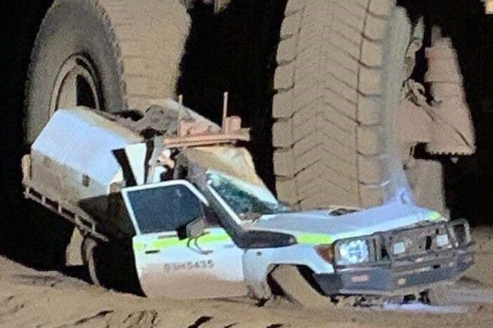 A white four wheel drive ute is crushed beneath the tyre of a large yellow dump truck