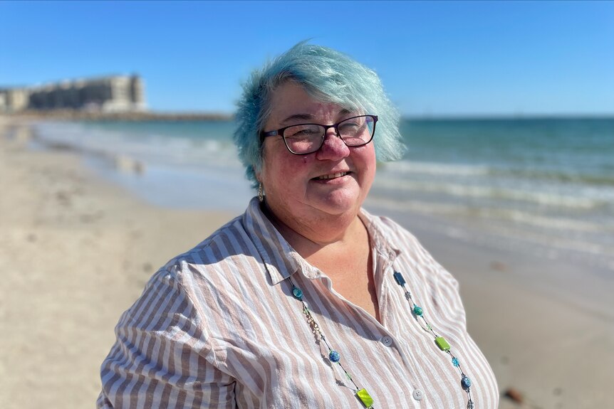 Белая женщина улыбается в камеру, у нее короткие синие волосы, очки и льняная рубашка.  Она стоит на пляже. 