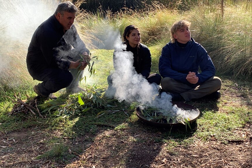 An Indigenous man holding gum leaves in front of a ceremonial fire with a young woman and teenage boy.