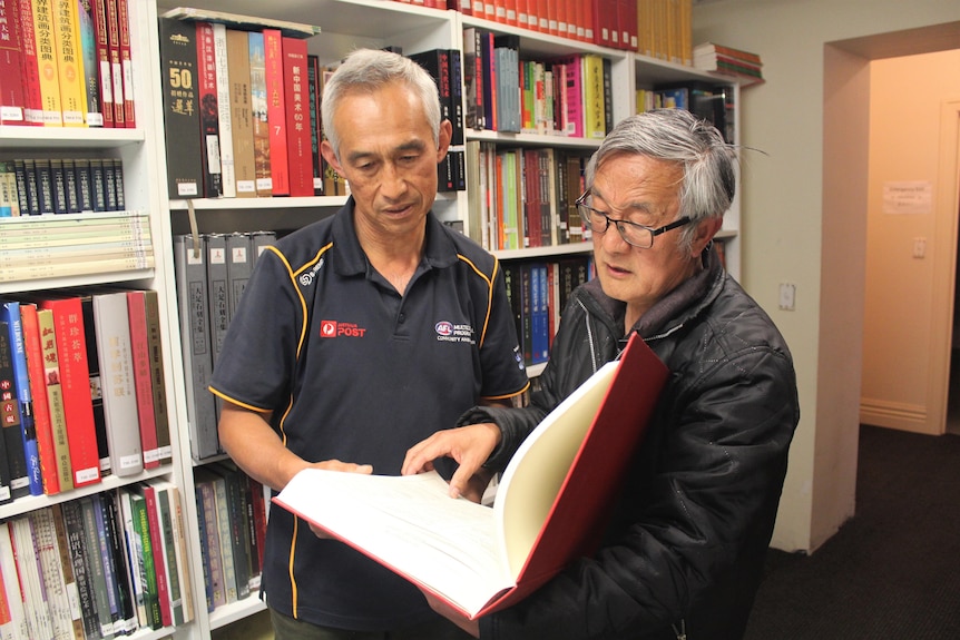 Two Chinese men look at a large Chinese book with book shelves behind them. 