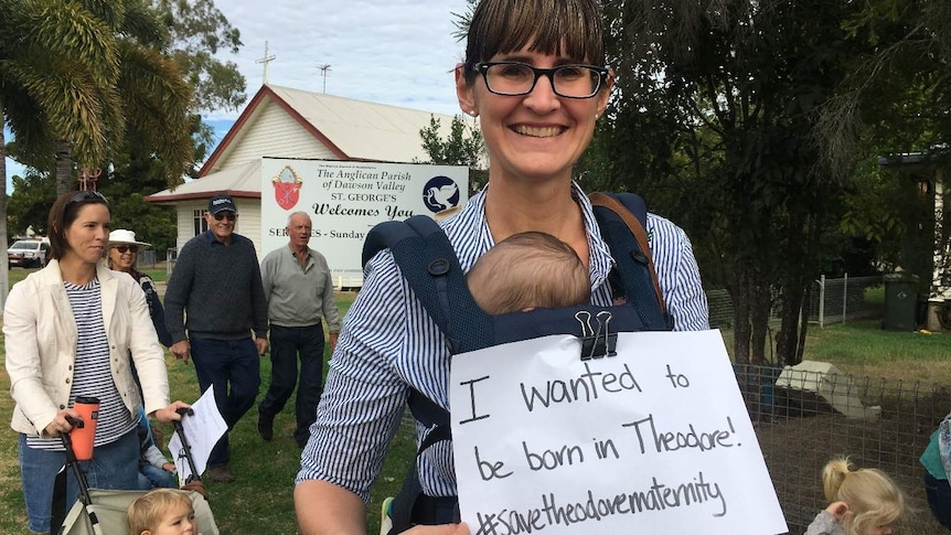 A woman holds her baby on her chest while holding a protest sign that reads "I wanted to be born in Theodore"