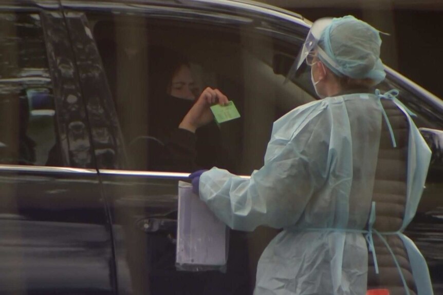 A woman being tested for coronavirus in a dark car as a health worker dressed in PPE looks on.