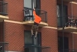 A man dressed in an orange top hanging off a black rail of a brick apartment building