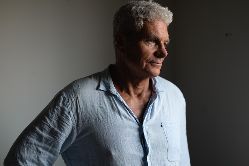 A man standing in a room looks to his side, with a serious and focused expression. Part of his face and the room is in shadow.