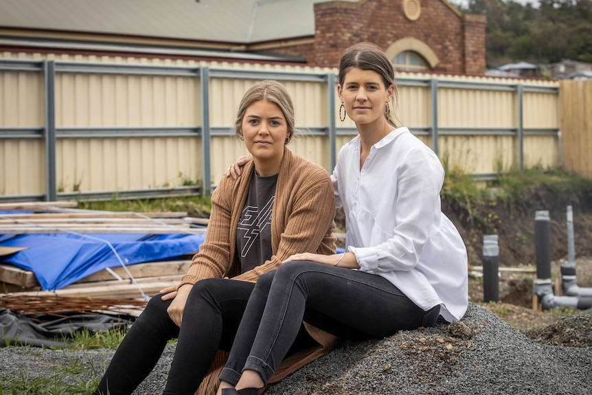 Two women sit on a pile of gravel at a building site.