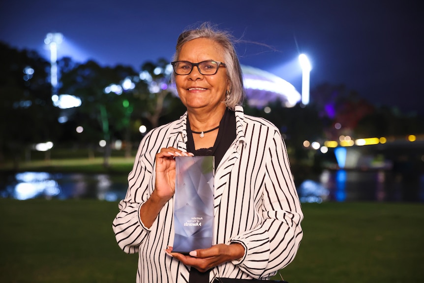 Woman smiling holds trophy at night outside exhibition centre.