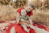 A woman sits with a baby on a rug