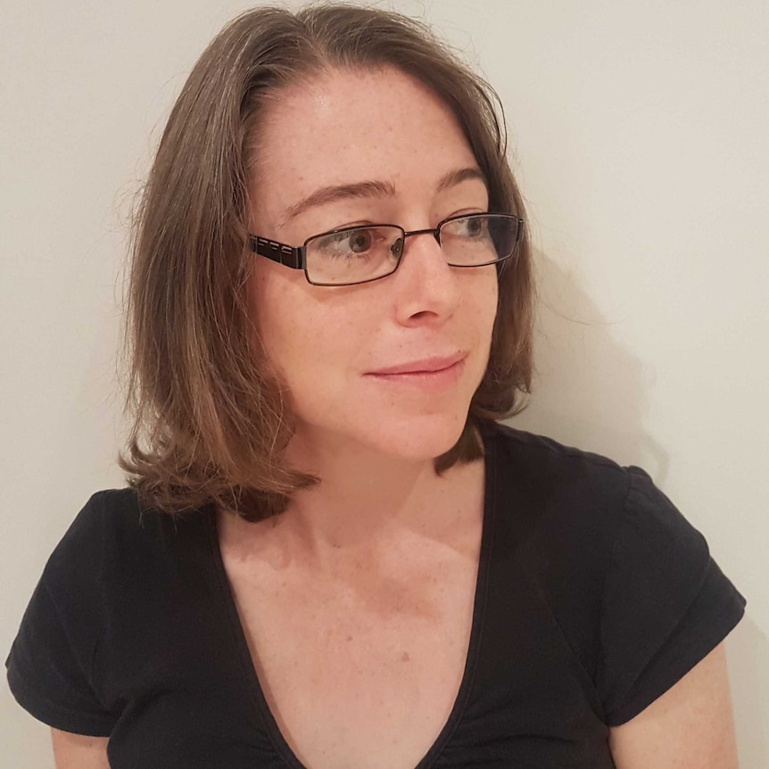 A close up of a woman standing against a white wall, wearing a black t-shirt and black-framed glasses.