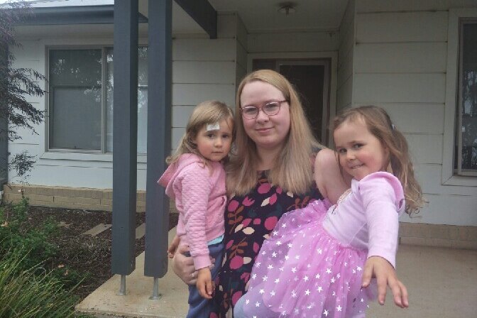 Teagan Sloman smiles as she stands in front of a house holding Zara on her left and Bree on her right.