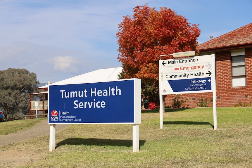 A blue Tumut Health Service sign outside a red brick building and other directional signs