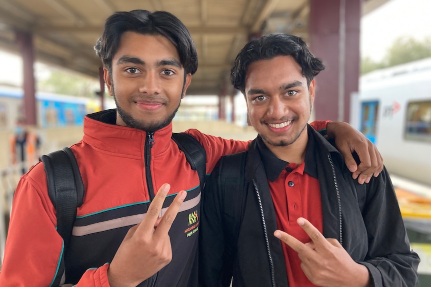 Hamdaan and Daniyal stand with their arm around one another smiling at a train station.
