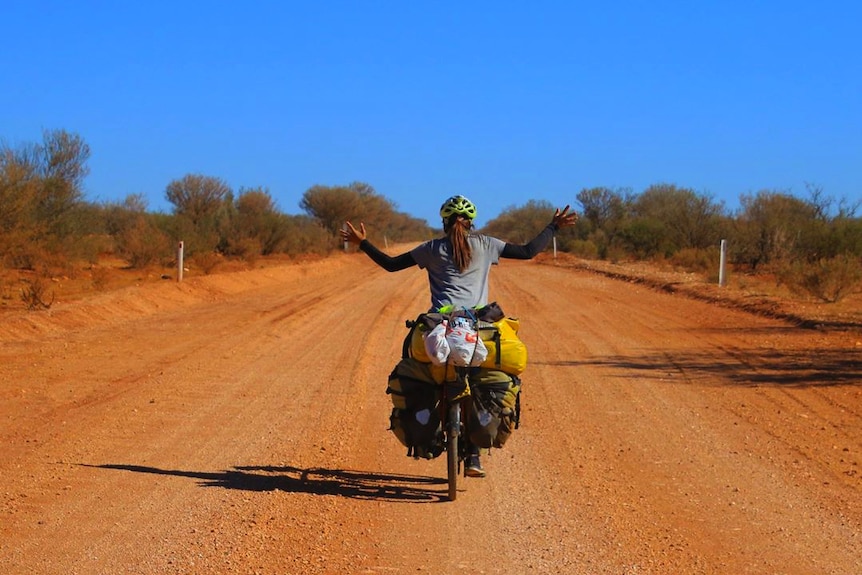 A woman, seen from behind, cycling down an outback road on heavily-laden bike.