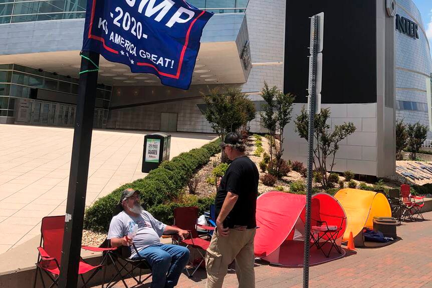 Two men on camping chairs outside a convention centre with a Trump flag flying