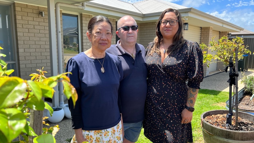 Two women stand on either side of a man on the front grass outside of a beige-brick home