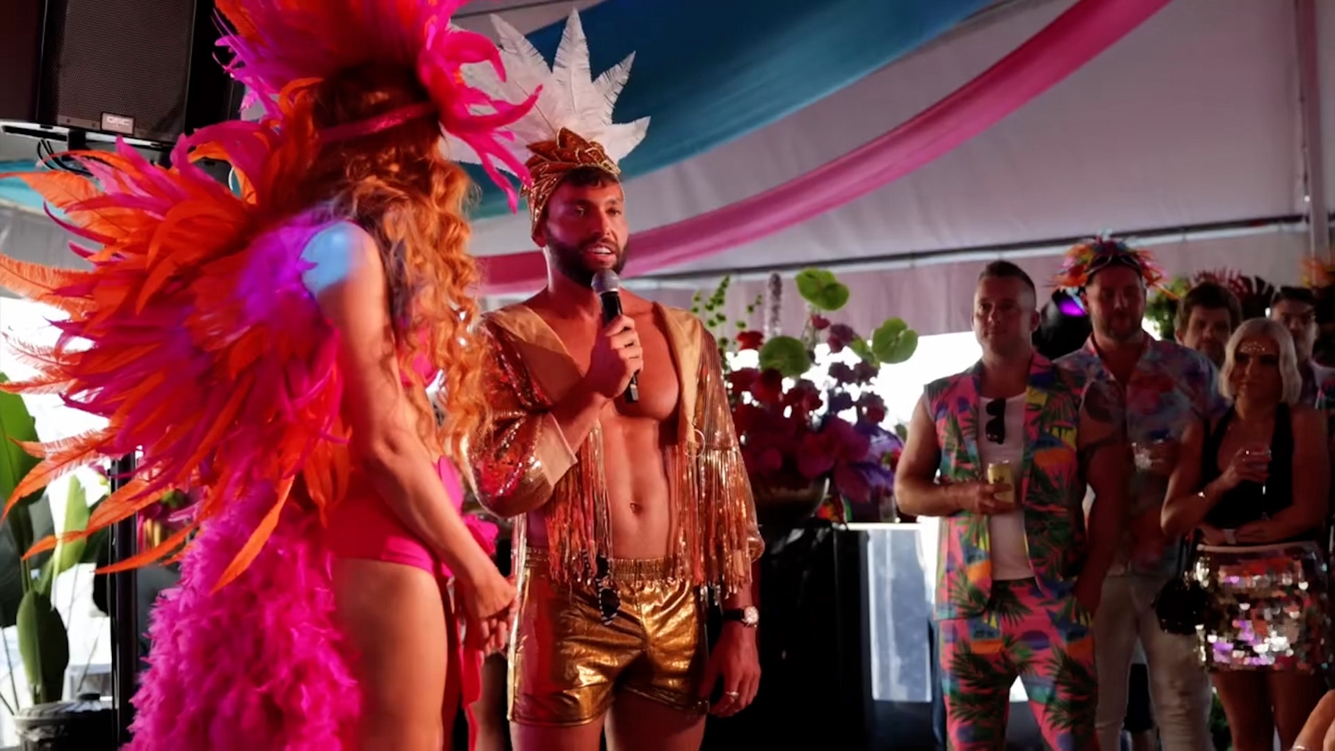 a man wearing gold shorts and a decorative headpiece speaks into a microphone as a carnival dancer and party guests look on