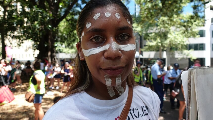 A young woman with face paint poses for a picture in front of a large crowd of protesters.