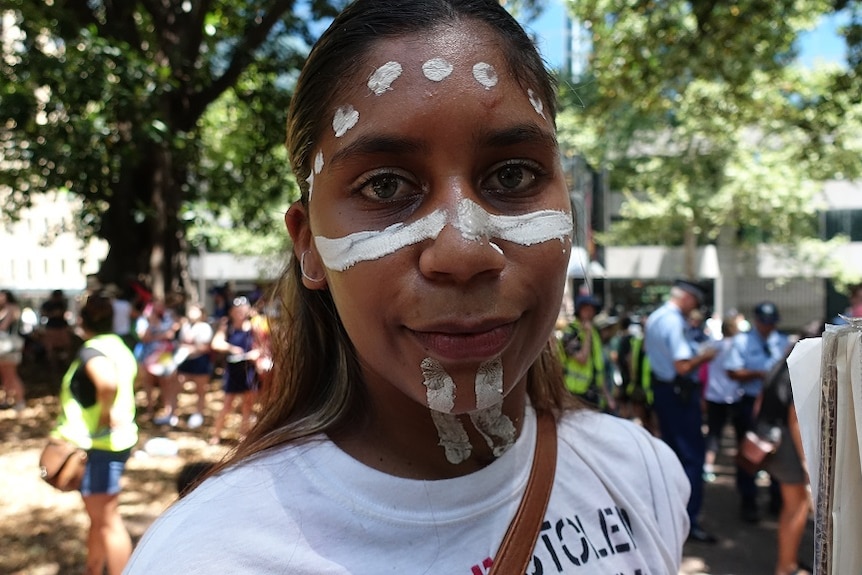 A young woman with face paint poses for a picture in front of a large crowd of protesters.
