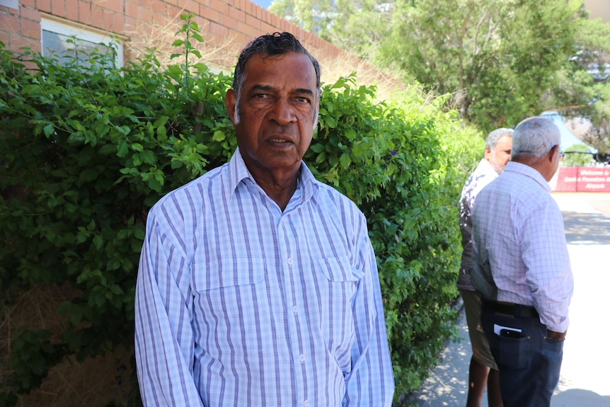 A First Nations man stands in from of a red brick building surrounded by a green shrub.