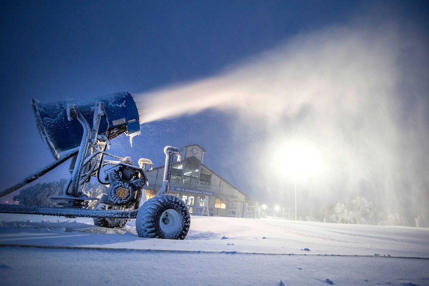 A snowmaker blows snow into the air on a ski slope, with ski and snowboard school in the background.