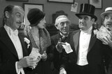 Several well dressed people hold up money and look at a man in a top hat and tuxedo who is frowning in a film still.