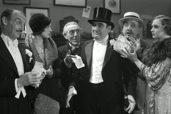 Several well dressed people hold up money and look at a man in a top hat and tuxedo who is frowning in a film still.