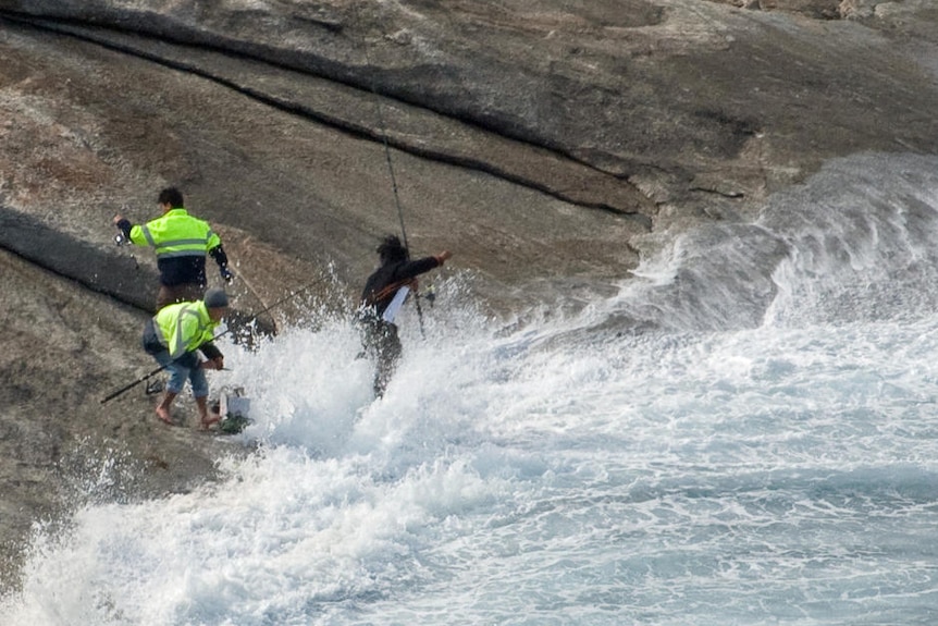 Huge wave washes onto fisherman on rocks that slope down into the sea