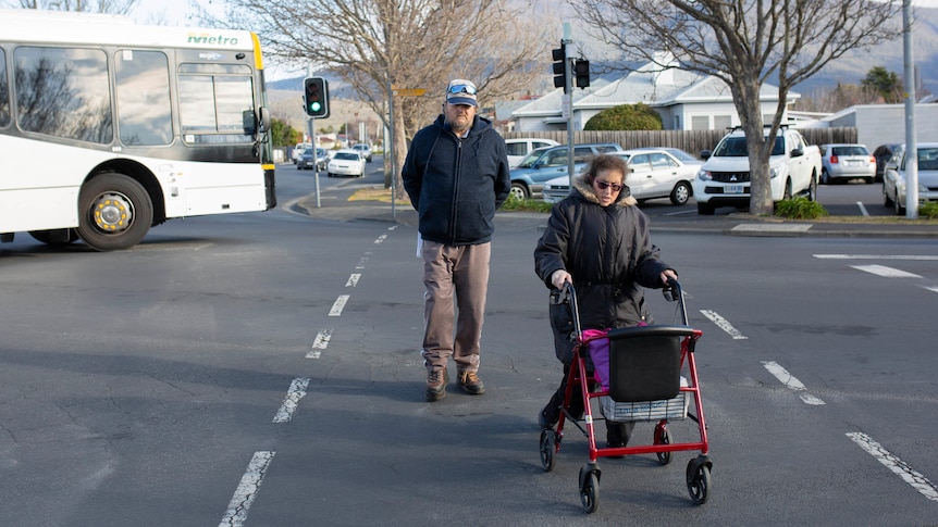 A woman uses a walking frame to cross the road with her father walking behind her.