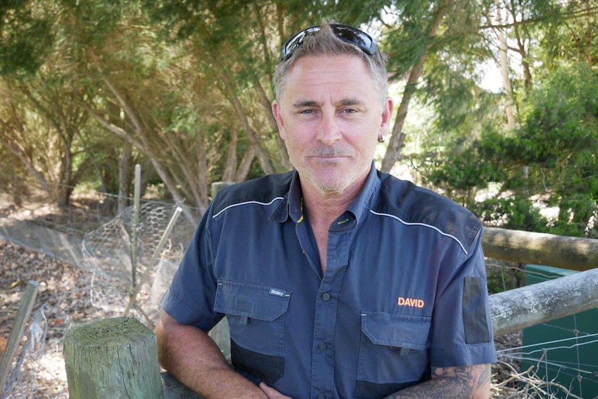 A picture of a man in a blue shirt leaning on a wooden fence post facing the camera.