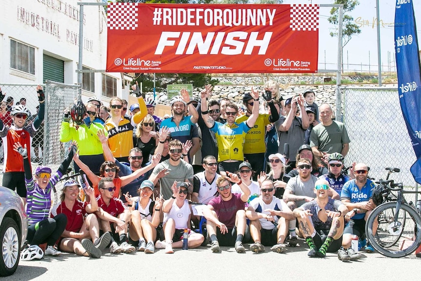 A large group of cyclists pose for a celebratory photo under a red banner that reads "Ride for Quinny finish".