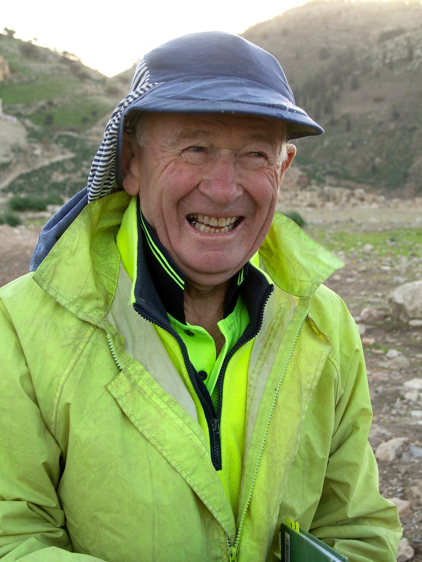 A man wearing a hat and a high visibility coat smiles with rocky hills in the background.