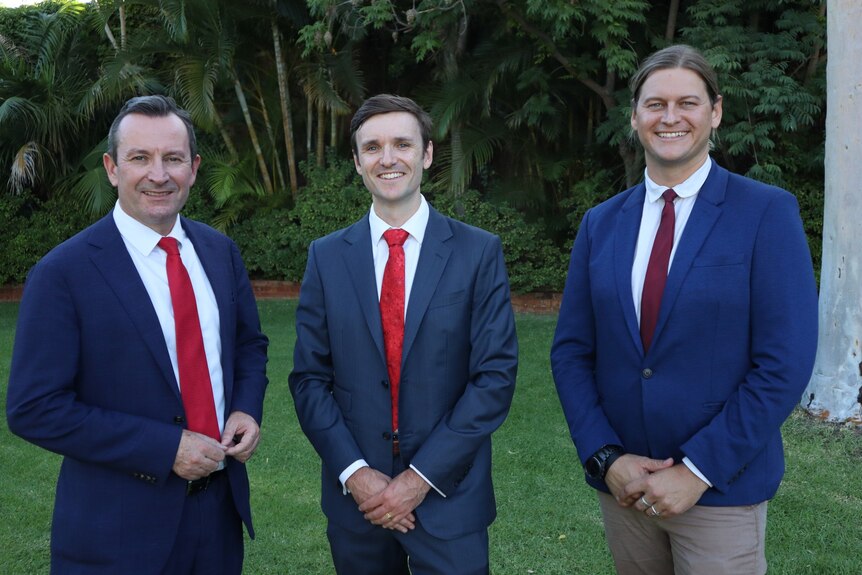 Mark McGowan, Yannick Spencer, and Stuart Aubrey stand in front of green gardens wearing blue suits, white shirts and red ties.