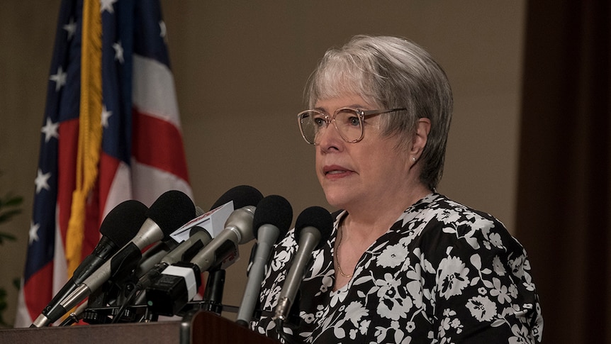 A woman with short grey hair, glasses and monochrome floral shirt stands at podium with several microphones near American flag.