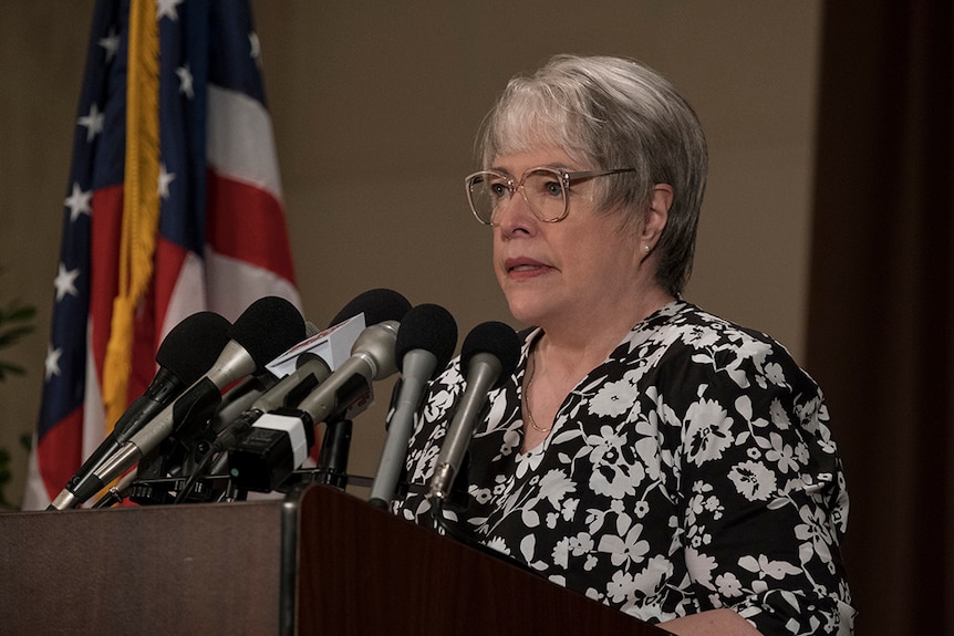 A woman with short grey hair, glasses and monochrome floral shirt stands at podium with several microphones near American flag.