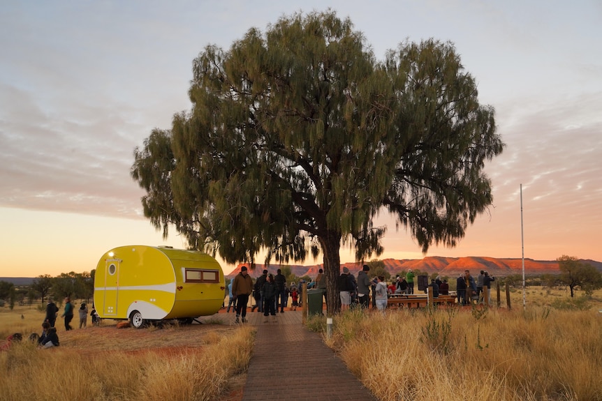 A yellow caravan sits under a huge tree, with a sillhouetted crowd facing Uluru in the distance, and sun setting behind it.
