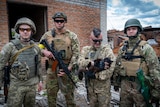 Four soldiers in army fatigues stand outside a brick home, one with a mohawk smiles adoringly at a black cat in his arms